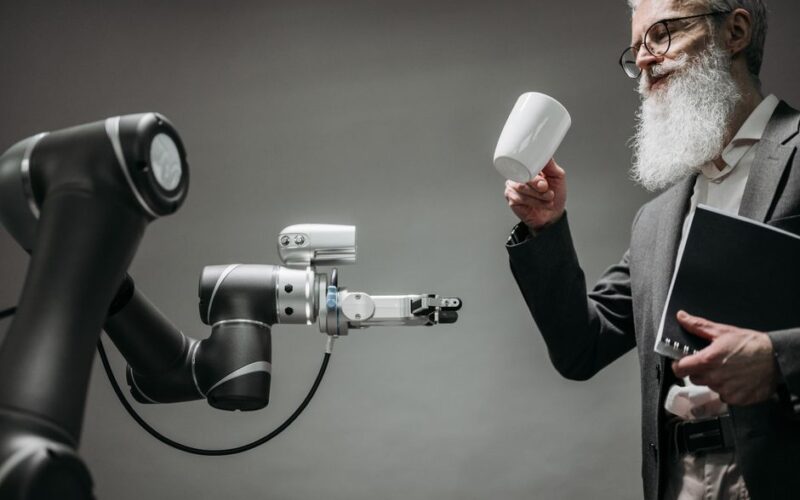 A bearded engineer holds a ceramic cup while a robotic arm assists, showcasing modern technology.