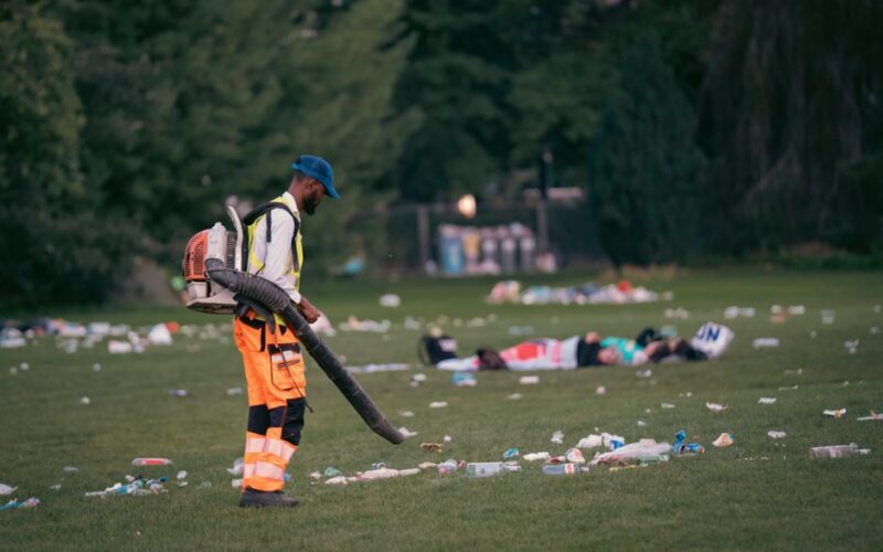Cleaning staff with leaf blower managing trash in a park after a public event.