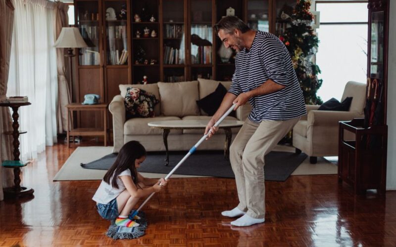 A playful family moment as a father and daughter mop the living room floor together, enjoying quality time.