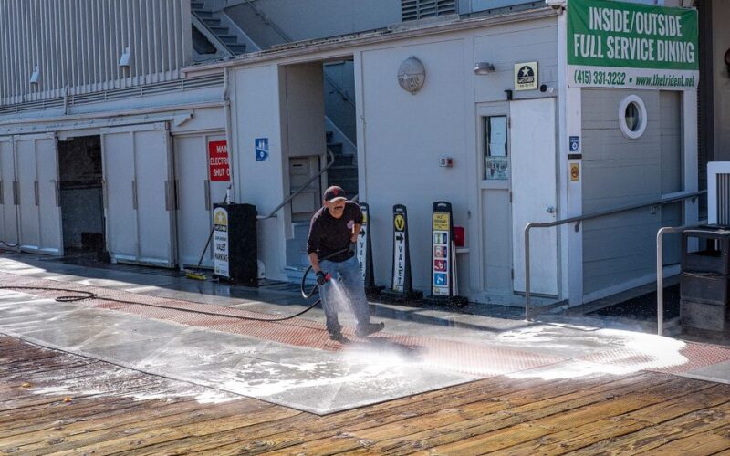 A worker uses a pressure washer to clean a sidewalk outside a dining facility.