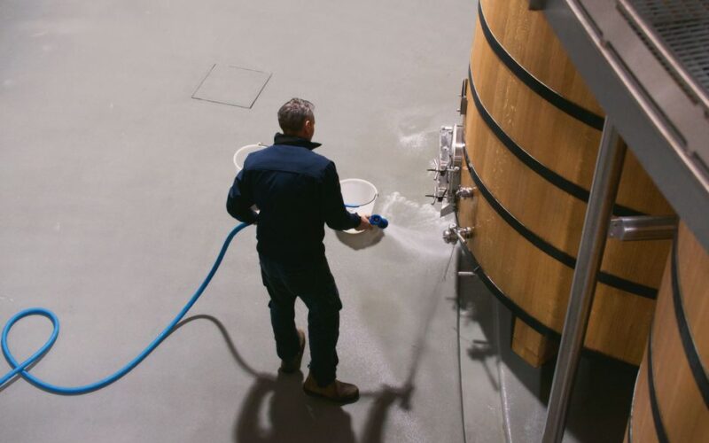 A winemaker cleans oak barrels in a Pauillac wine cellar in France, using a hose to maintain hygiene.