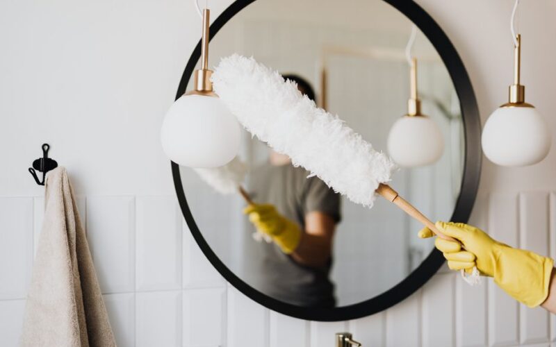 Crop anonymous female housewife in rubber gloves dusting modern lamp with soft brush against mirror while cleaning stylish bathroom