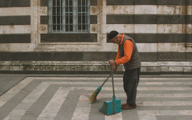 Elderly man cleaning a striped courtyard with broom and dustpan outside a historic building.