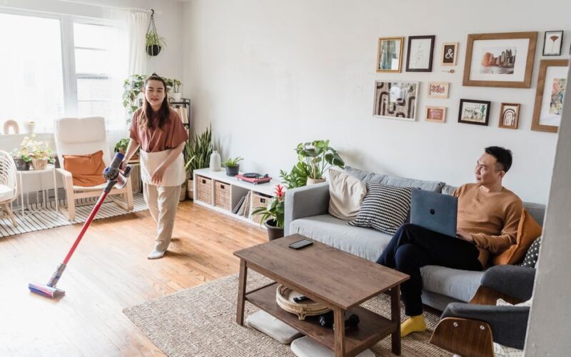 A man and woman indoors, one cleaning, the other using a laptop, in a modern living room.