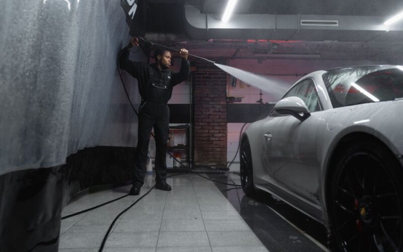 A man uses a pressure washer to clean a white car in an indoor car wash.