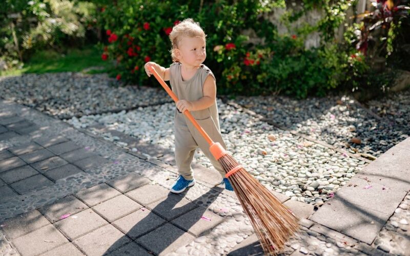 A cute child sweeping a garden pathway with a broom under sunny weather.