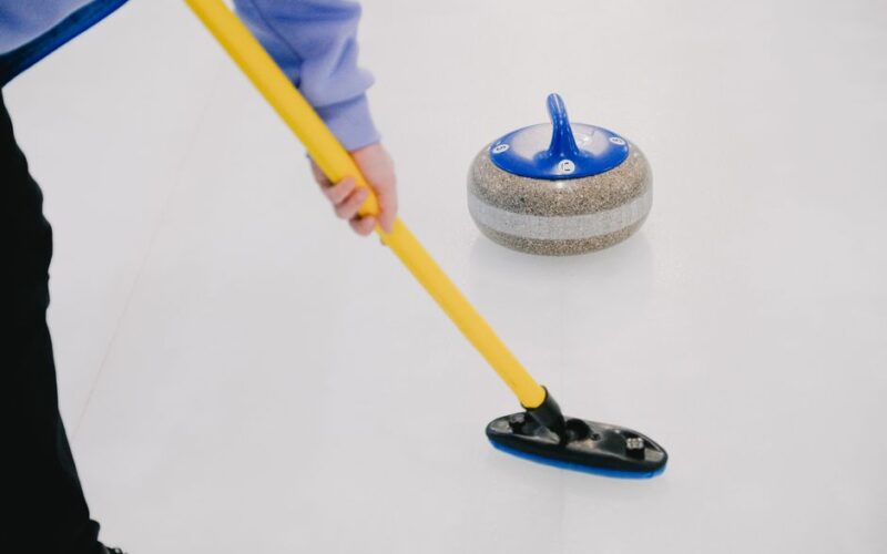 Crop anonymous sportsman with special broom sweeping ice sheet in front of sliding curling granite stone while playing sportive game