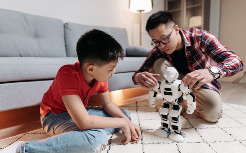 A father and son engage with a humanoid robot on a living room carpet, fostering creativity.