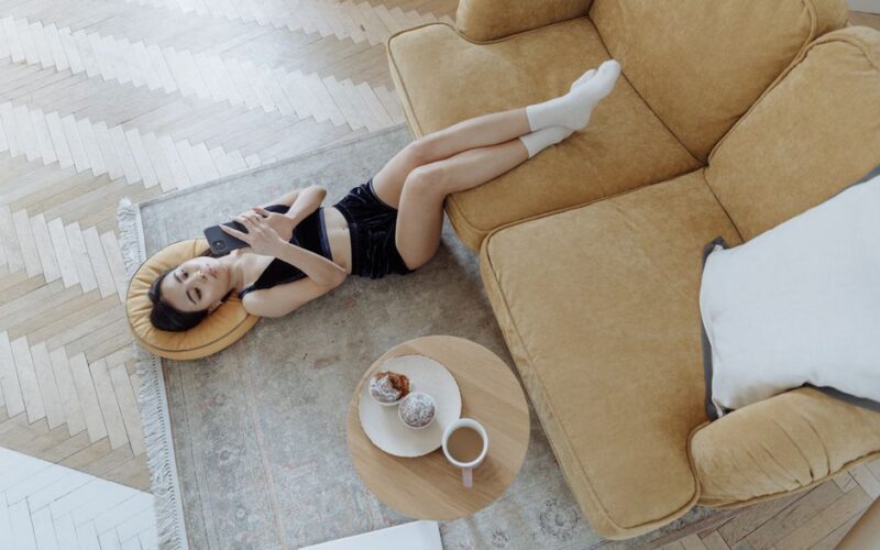 Woman lying on carpet using cellphone with snack and coffee nearby, enjoying a relaxing moment indoors.