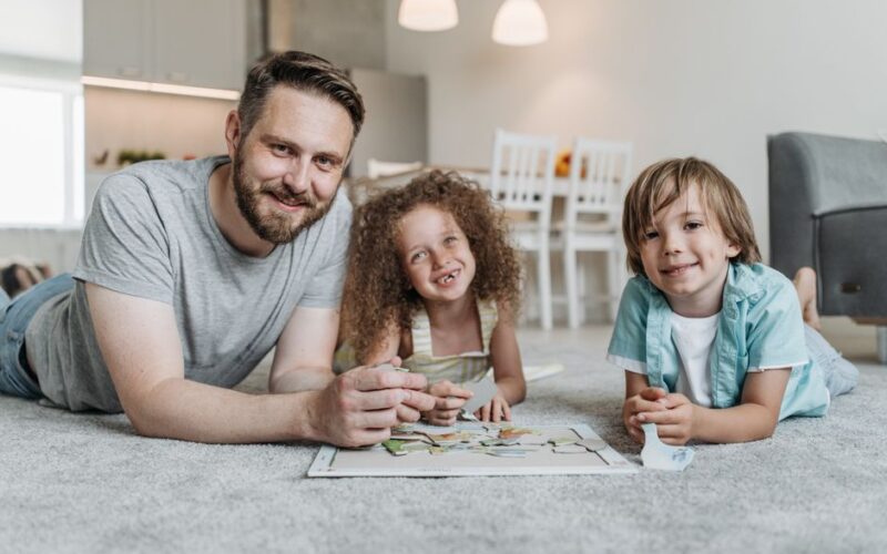Father and two children smiling while solving a puzzle together on a soft carpet indoors.