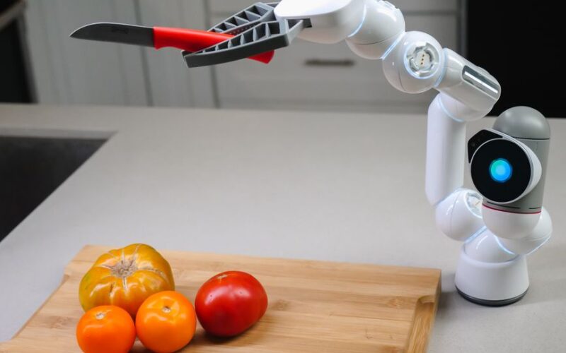 A futuristic robotic arm with a knife preparing colorful tomatoes in a modern kitchen.