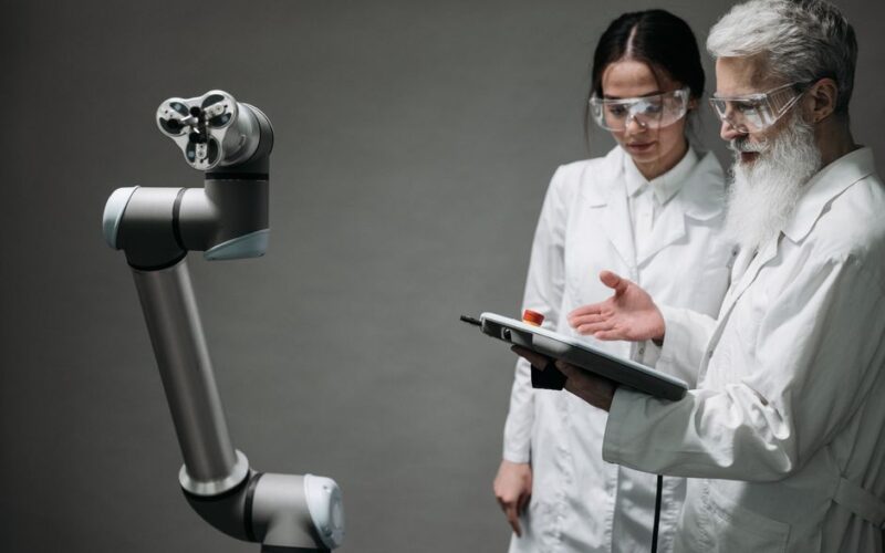 Researchers in lab coats and safety glasses engaging with a robotic arm in a lab setting.