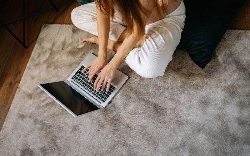 A woman in comfortable attire typing on a laptop while seated on a soft carpet, creating a warm work-from-home atmosphere.