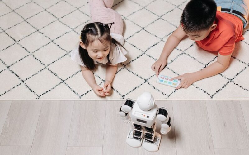 Two children control a playing robot with a mobile device indoors, engaging in technology.