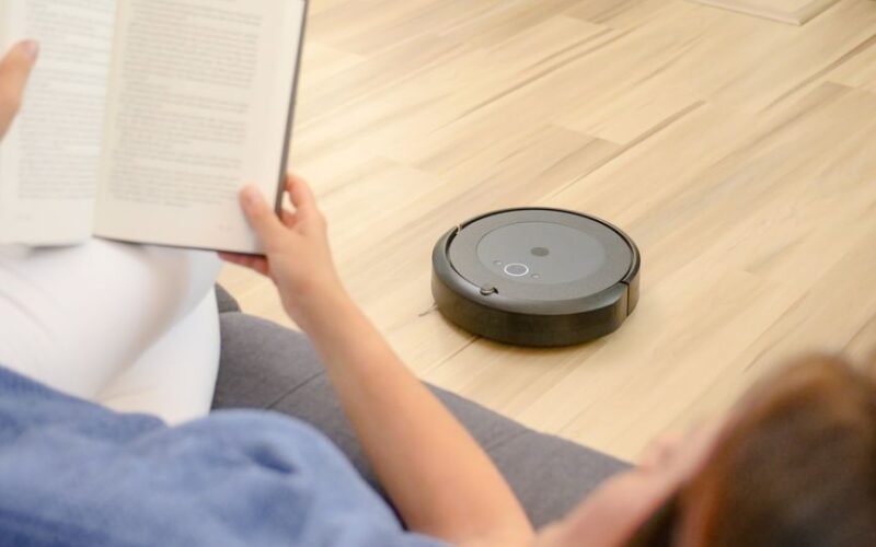 A woman relaxes on a sofa reading while a robotic vacuum navigates the floor.