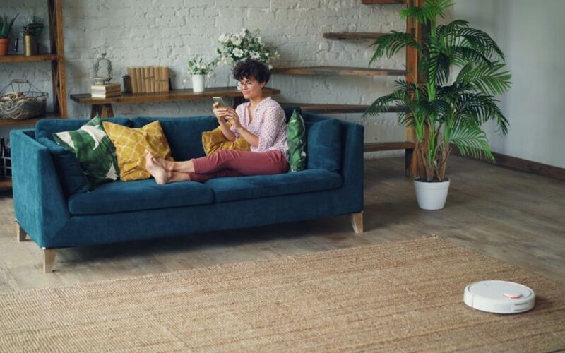 Woman relaxing on a stylish blue sofa at home, using a smartphone with a robotic vacuum on the floor.