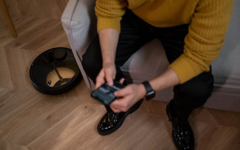Man in yellow sweater using remote to control robotic vacuum on wooden floor.