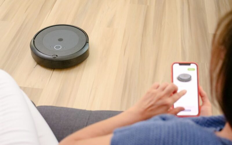 A person uses a smartphone app to control a robotic vacuum on a hardwood floor, showcasing smart home technology.