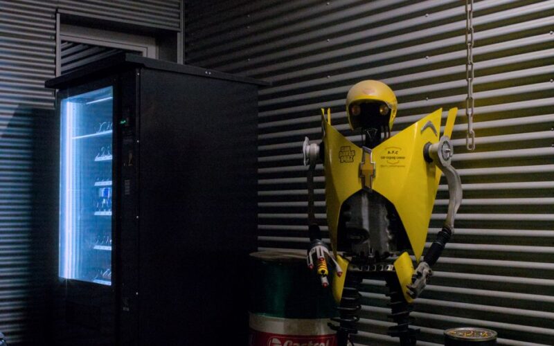 A dimly lit industrial room features a vending machine beside a quirky robot sculpture.