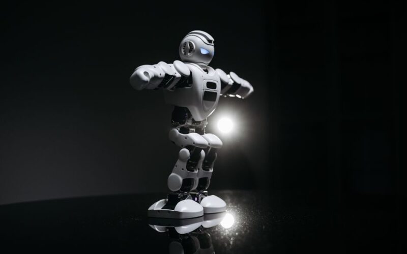 White humanoid robot with glowing eyes standing on a reflective black surface in studio lighting.