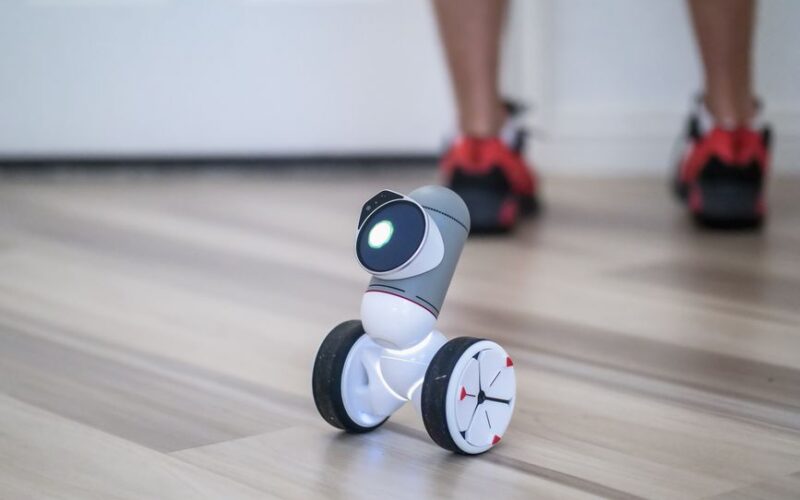A close-up of a modern wheeled robot on a wooden floor with legs in the background.