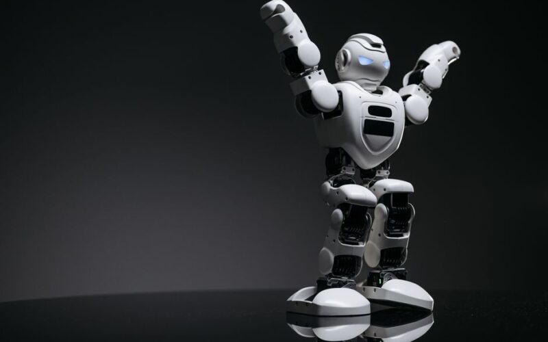 Close-up studio shot of a white robot toy with LED eyes raised in victory on a gray background.