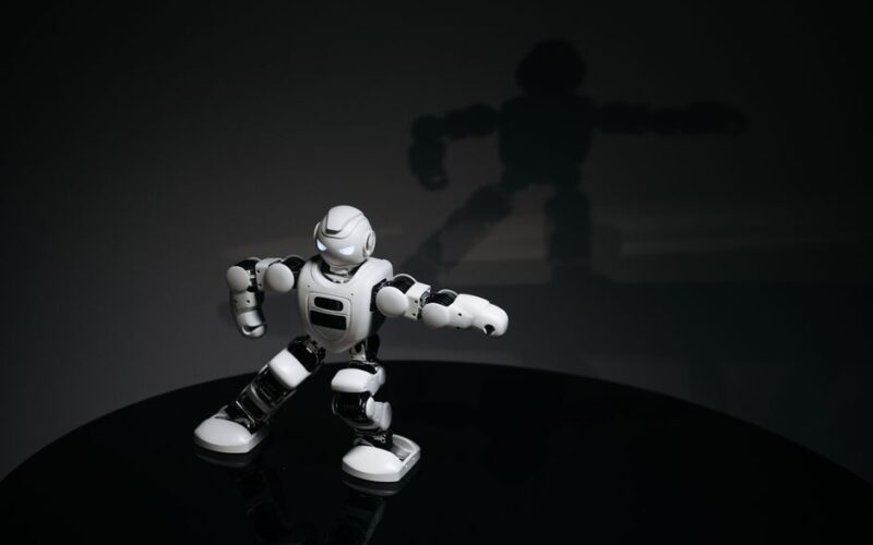 A futuristic white toy robot striking a pose in a dimly lit studio with a shadow cast behind it.