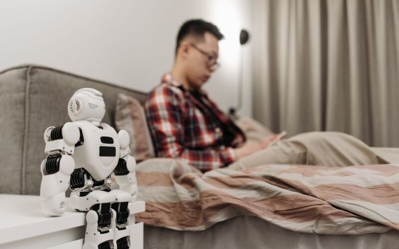 A modern bedroom featuring a robot on a bedside table and a man sitting on a bed.