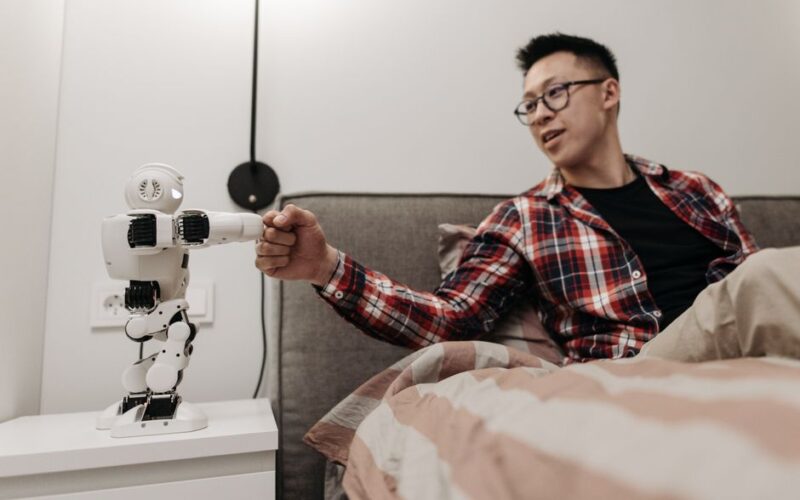 Asian man sitting on bed shaking hands with a robot in a modern bedroom.