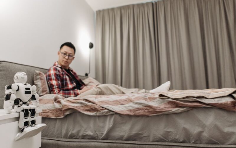 Asian man sitting in bedroom, observing futuristic robot on nightstand.