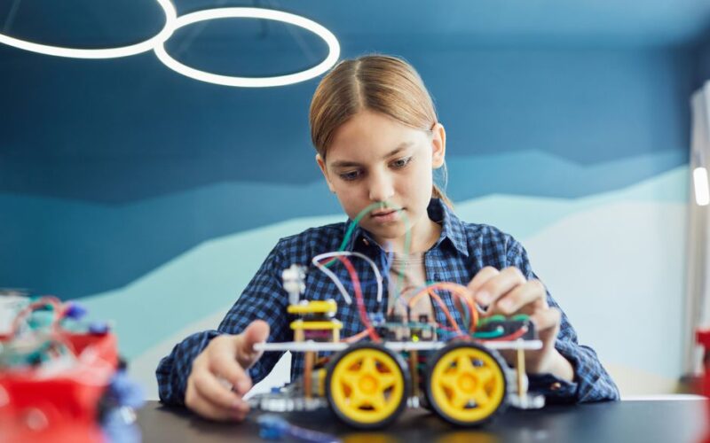 Teenage girl working on a robotic car project with wires and wheels inside a classroom.