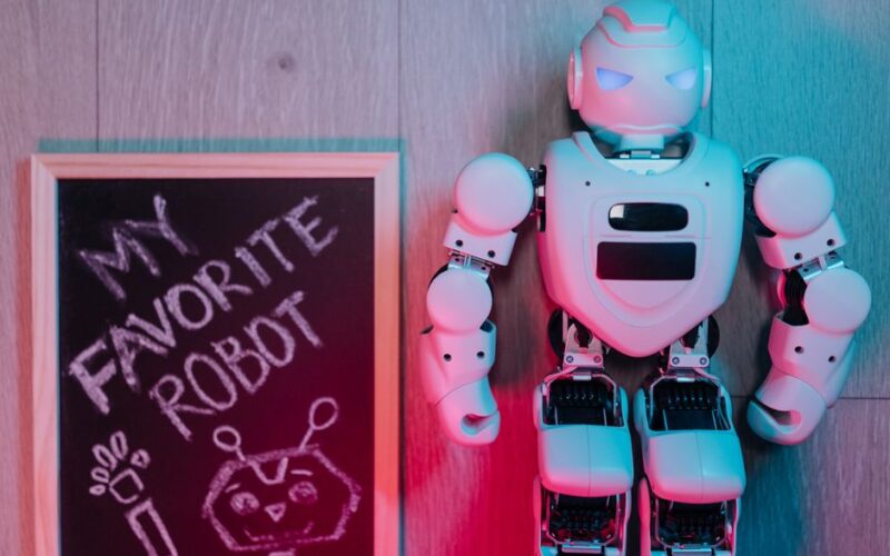 A futuristic toy robot standing beside a chalkboard on wooden flooring, highlighted by colored lighting.