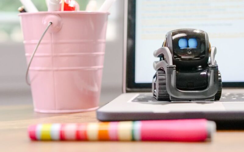 A small robot sits on a laptop keyboard beside a pink pen holder, symbolizing technology and innovation.