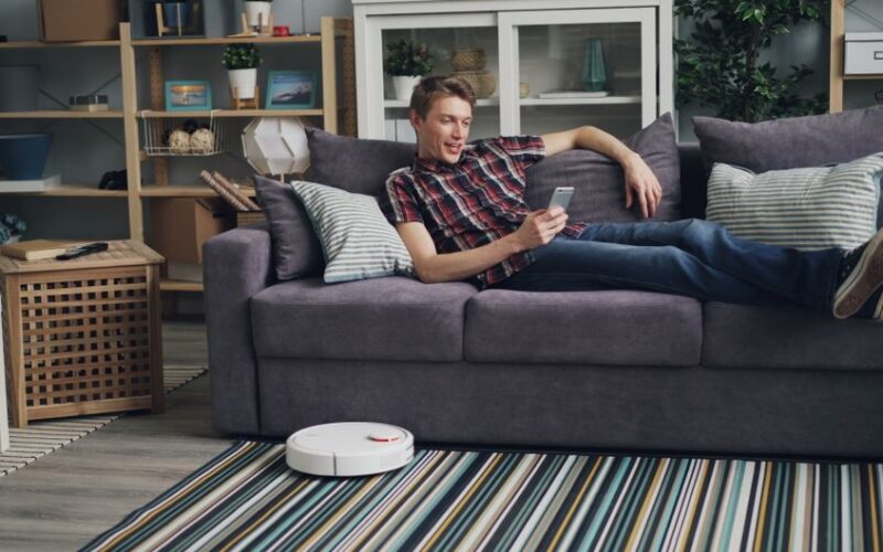 Man lying on a sofa using a smartphone while a robot vacuum cleans the floor in a cozy living room.