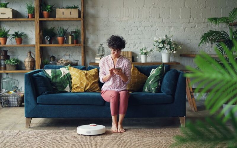 Woman sits on sofa using smartphone while robotic vacuum cleans living room floor.