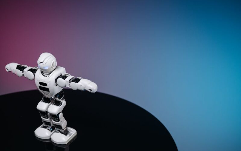 A white humanoid toy robot standing on a reflective black surface in a studio setting with a blue and pink gradient background.