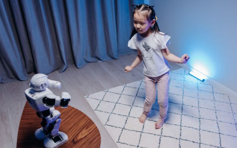A young girl joyfully dances with a robot on a rug in a modern, softly lit room.