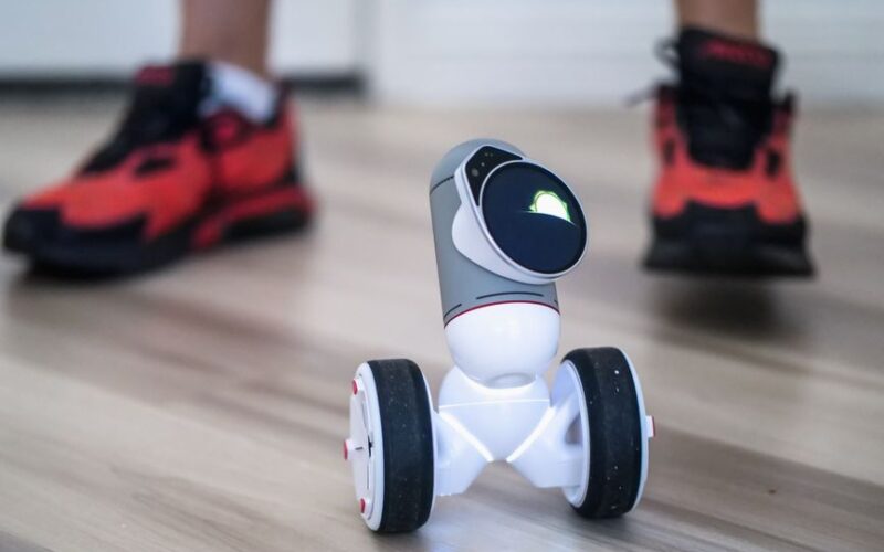 Close-up of a two-wheeled mini robot on a wooden floor with sneakers in background.