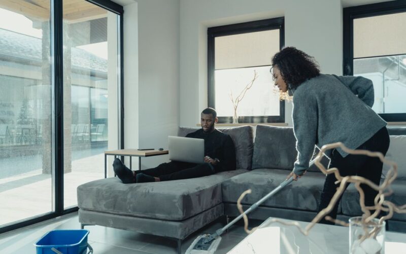 A couple in a modern living room, one using a laptop, the other cleaning.