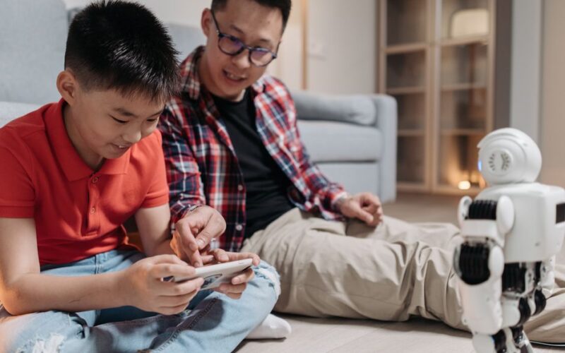 A father and his son sit on the floor, interacting playfully with a robot using a smartphone.