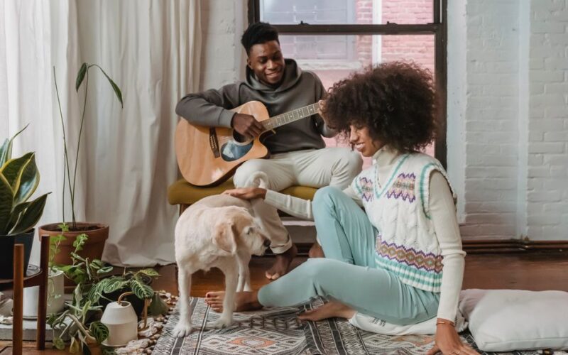 Full body of cheerful African American boyfriend playing guitar near ethnic happy girlfriend with dog on rug in cozy lounge