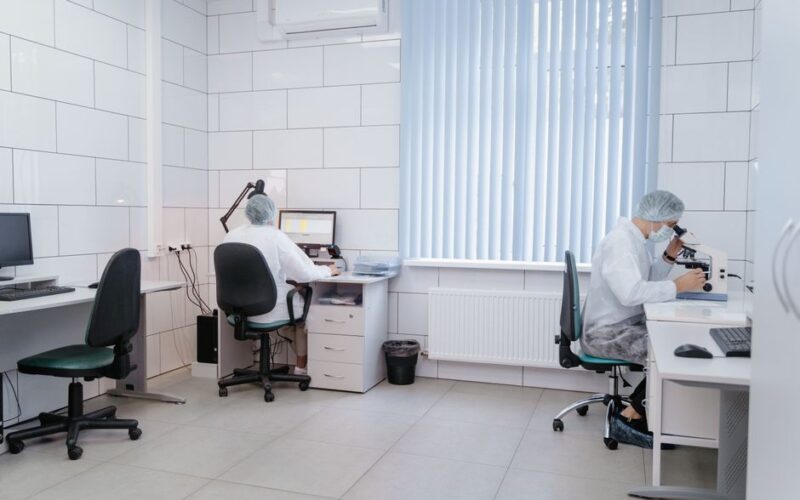 Two laboratory technicians working with computers and microscopes in a clean, modern lab.