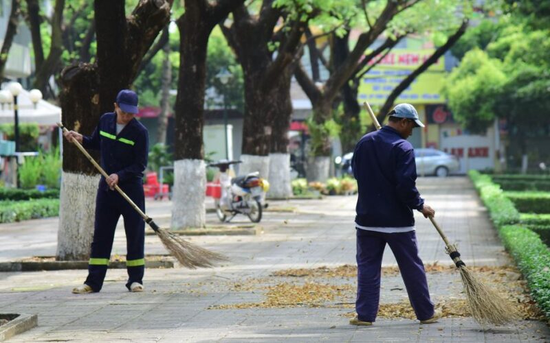 Two workers sweeping a tree-lined sidewalk, maintaining clean urban environment.