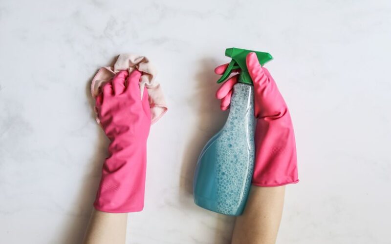 A pair of hands in pink gloves are cleaning with a spray bottle on a white background.