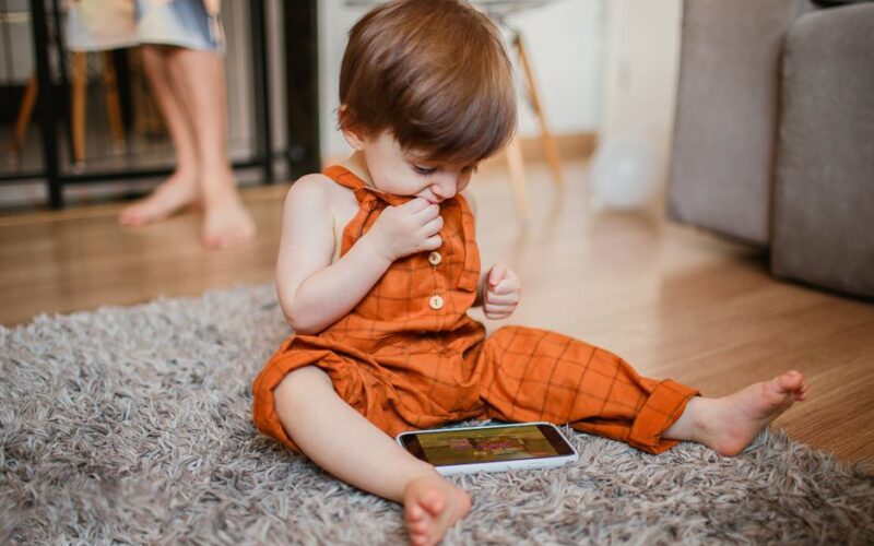 Cute toddler in orange outfit plays with smartphone on carpet indoors.