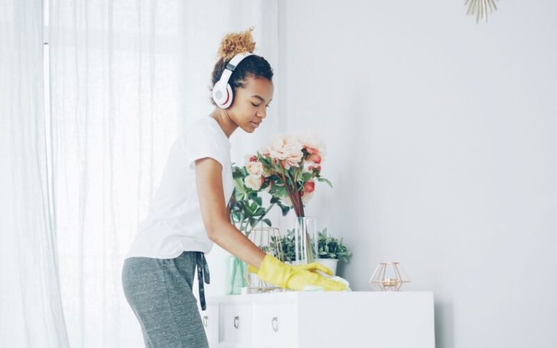 Woman in white shirt cleans table while listening to music with headphones. Bright indoor scene.