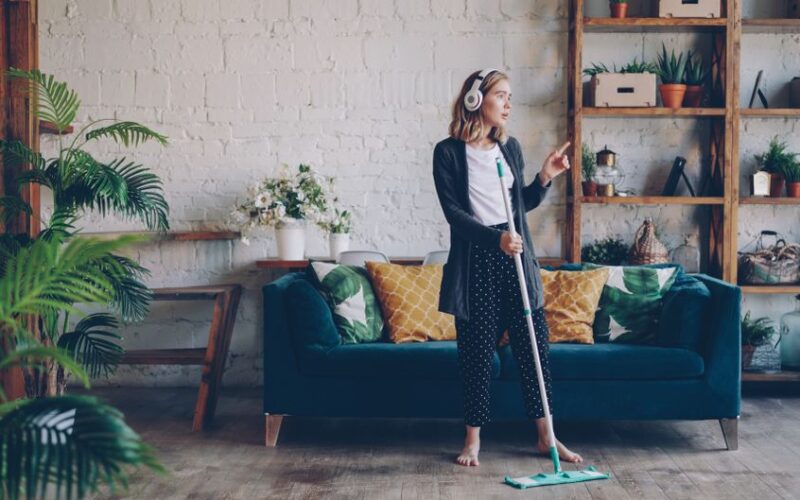 Cheerful woman with headphones mopping floor in cozy living room interior.