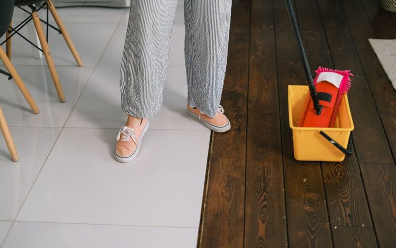 Crop anonymous woman in gumshoes and pants dipping mop into bucket with water on parquet in house