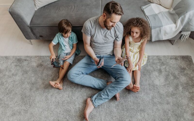 Father and children playing with toys on a gray carpet in a cozy living room from above.