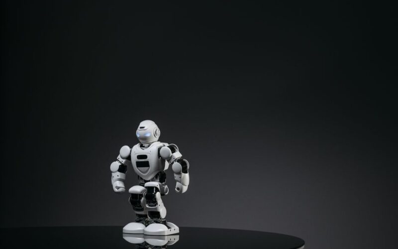 A small white robot stands on a reflective surface in a dark studio setting, showcasing modern technology.
